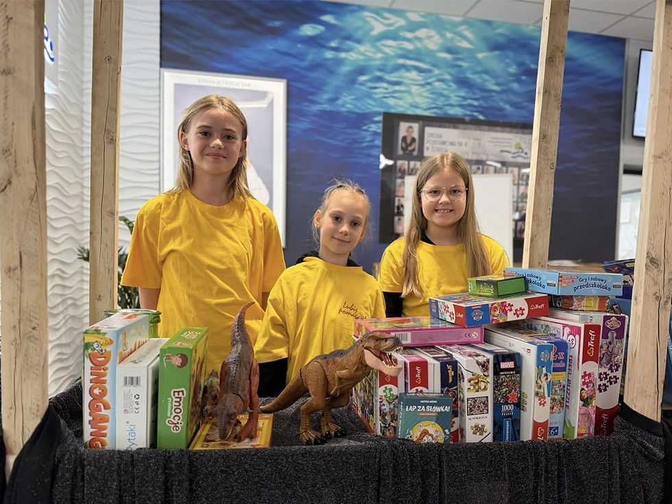 Trzy dziewczynki w żółtych koszulkach stojące przy straganie zastawionym grami, układankami i zabawkami