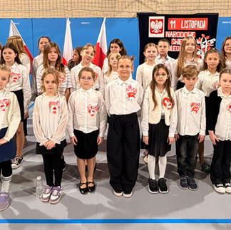 Grupa dzieci w strojach galowych stoi na sali gimnastycznej przed dekoracją z napisem „11 listopada – Narodowe Święto Niepodległości”.