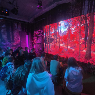 Uczniowie siedzą przed dużym ekranem z baśniowym krajobrazem w czerwonych barwach, oglądając multimedialną prezentację.