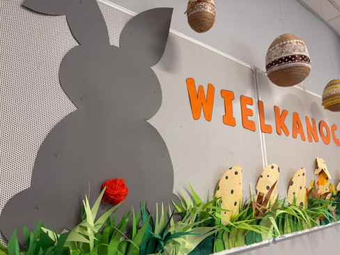 Dekoracja na ściennej tablicy - szary zajączek i kolorowe pisanki na trawie z papieru, nad nimi napis: Wielkanoc