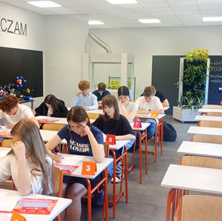 Uczniowie siedzą w ławkach w sali lekcyjnej pochyleni nad kartkami.