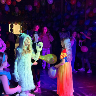 Dzieci w strojach karnawałowych tańczące na sali udekorowanej balonami, w kolorowym świetle dyskotekowych reflektorów.