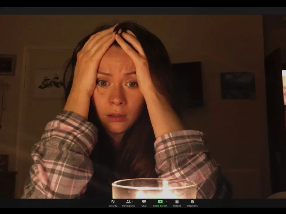 An anxious woman on a video call holds her head in a dim room, lit by a candle; Zoom toolbar icons appear at the bottom.
