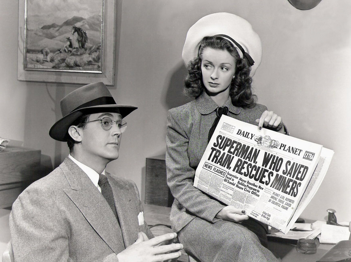 Actor Kirk Alyn, seated in a suit and glasses, gestures as actress Noel Neill, wearing a hat, shows him a Daily Planet newspaper headlined "Superman Rescues Miners."