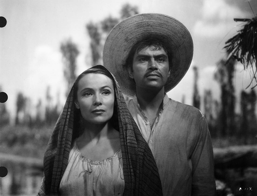 The black-and-white image features Delores del Río and Pedro Armendáriz standing outdoors in "María Candelaria" (1944). Del Río is on the left; her long hair is partially covered by a patterned shawl draped over her head. She wears a light-colored blouse and looks forward with a serene expression. Armendáriz is on the right; he has dark hair and a mustache, wearing a wide-brimmed straw hat and a light shirt. He looks slightly off-camera with a determined expression. The background is blurred, showing tall, slender trees and a cloudy sky, indicating a rural, possibly agricultural setting.