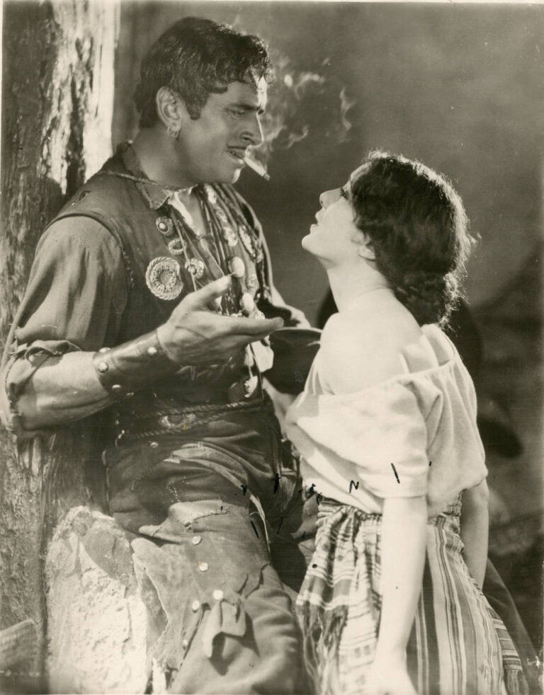 Man, Douglass Fairbanks, smoking, wearing an adorned vest, leans against a stone wall, facing a woman, Lupe Vélez, in a striped skirt and off-shoulder top. Intense, dramatic scene in The Gaucho (1927).