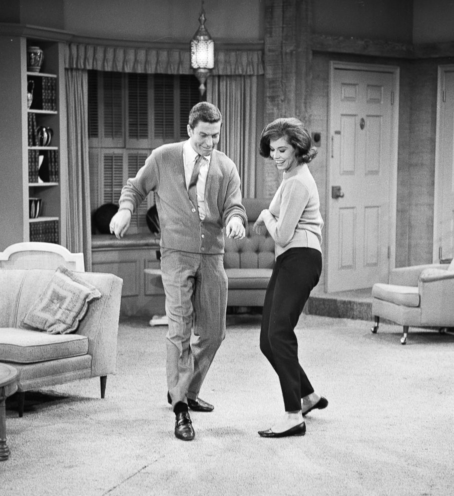 Dick Van Dyke and Mary Tyler Moore in the "Dick Van Dyke Show." The image is a black-and-white photograph of the two dancing in the show's living room set. Van Dyke, on the left, is wearing a light-colored cardigan over a shirt and tie, with trousers and dress shoes. Moore, on the right, is dressed in a sweater and slim pants with ballet flats. They are both smiling and appear to be enjoying themselves. The background shows a mid-century modern living room with a sofa, a bookshelf filled with various objects, and a hanging lamp. The floor is carpeted, and a small table with a cushion is visible in the foreground. Curtains cover the window behind the couch, and a door is visible to the right.