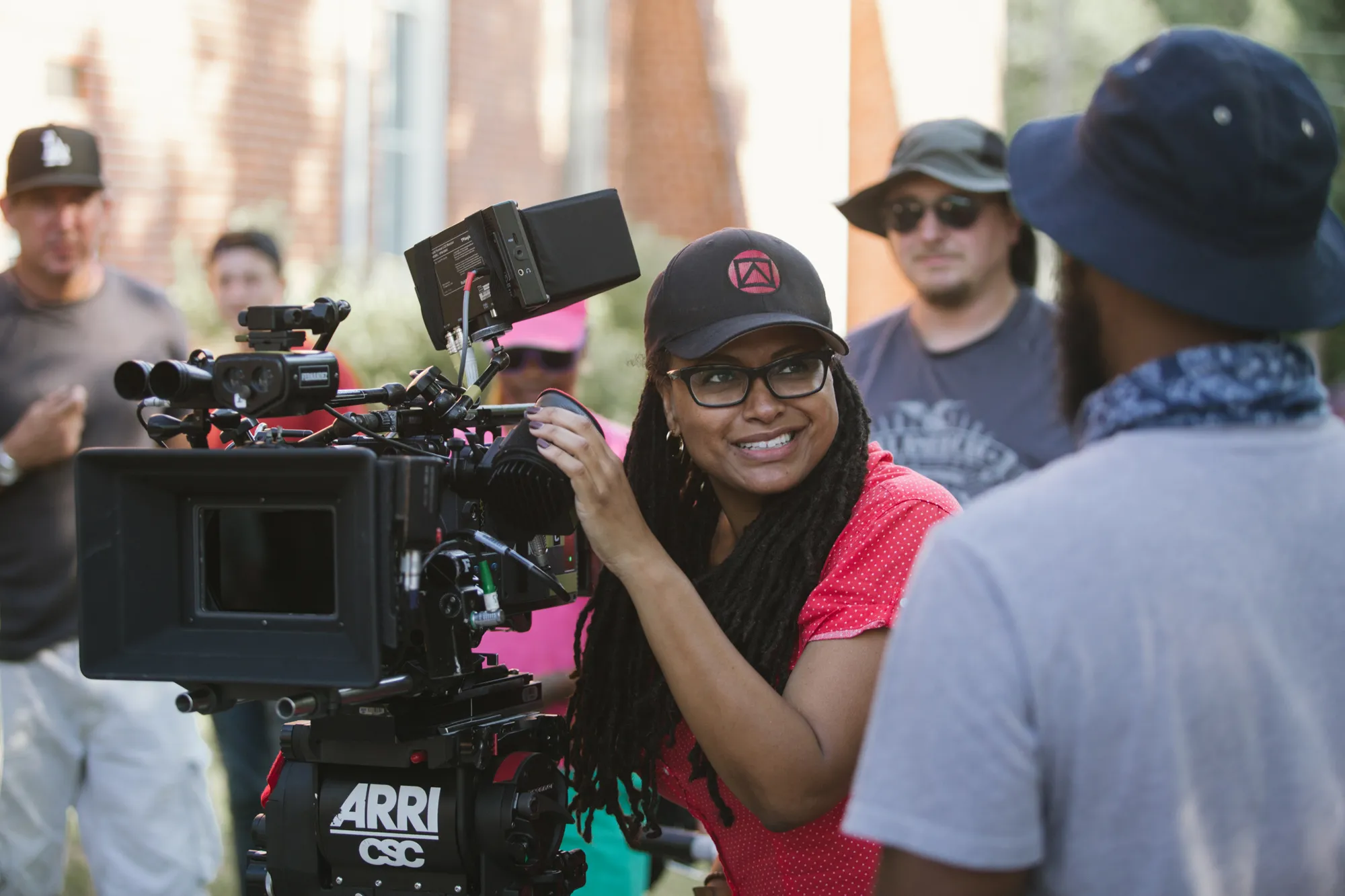 The image shows a film production scene outdoors with a focus on Ava DuVernay operating a large film camera. DuVernay, wearing a black cap with a red symbol and glasses, appears to be smiling and engaged. She has long dreadlocks and is wearing a red top. The camera is a professional ARRI model with various attachments. In the background, several people are visible, some wearing hats and casual clothing. The setting appears bright with soft natural light and a blurred background of greenery and a brick structure.