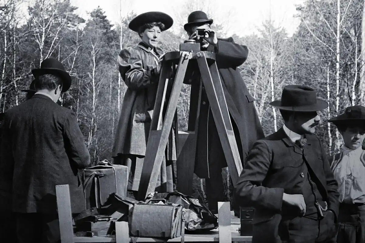 The black-and-white photo shows a group of people outside, gathered around a tripod-mounted camera. The main person is a woman, Alice Guy-Blaché, standing on a raised platform next to a man operating the camera. Both are dressed in early 20th-century clothes; Guy-Blaché wears a wide-brimmed hat and a long coat, while the man has a bowler hat and a heavy overcoat. Surrounding them are other people, including a man in a suit and hat, casually with his hand on his hip. In the background, leafless trees form a textured scene, indicating winter or early spring. Equipment and bags are scattered on the platform, showing a focus on photography or filming.