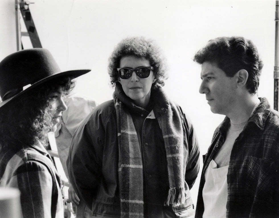 Three people converse outdoors on the set of the film Crossing Delancey (1988). On the left, Amy Irving is wearing a large hat and a scarf; in the middle, Joan Micklin Silver is wearing sunglasses and a scarf; on the right, Peter Riegert is wearing a dark coat. The mood is serious. Background is neutral.