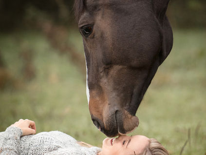 Eine Frau liegt im Gras und ihr Pferd berührt mit seinem Maul ihre Nasenspitze
