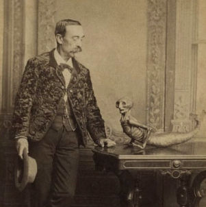Antique cabinet card photograph showing a gentleman in a patterned jacket looking at a taxidermy mermaid prop—a hoax made from a monkey and a fish—resting on an ornate wooden table. Printed text at the bottom reads "G. A. Garrett, Bloomington, Ill."