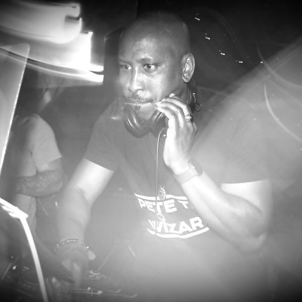DJ Pete the Wizard mixing music live at an event. A black and white action shot showing him focused on his DJ controller with headphones to his ear, wearing a branded Pete the Wizard T-shirt.