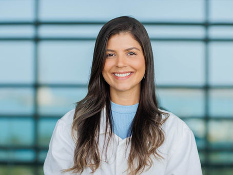 A professional headshot of Dr. Helena Villela smiling, wearing a white lab coat over a blue shirt, with a blurred modern architectural background.