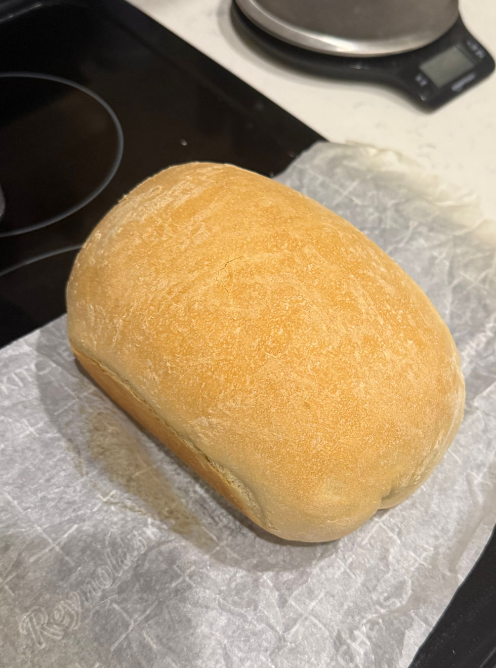 Golden loaf of bread on parchment paper, near a stovetop. Kitchen scale in the background. Warm and inviting atmosphere.