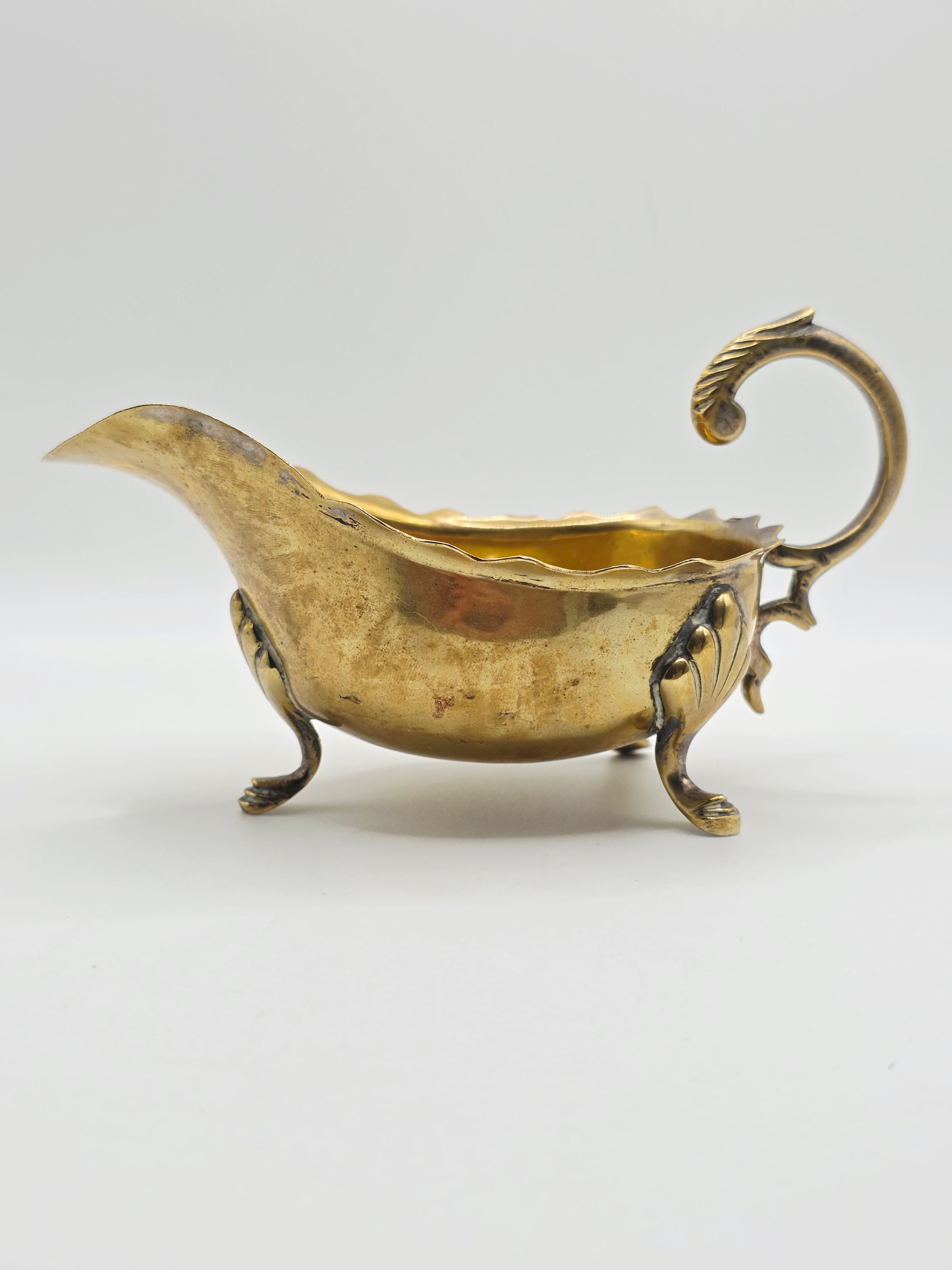 Side profile of vintage brass sauce boat with scrolled handle and shell pad feet.