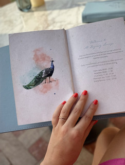 A hand with coral nail polish holding open a menu featuring a watercolor illustration of a peacock and Al Fayrooz Lounge’s operating hours.