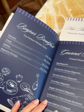 Hand holding open Le Fournil’s navy blue menu, showing the Parisian breakfast and gourmet baguette options on a marble table.