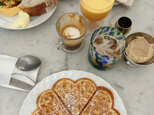 Plate featuring a heart-shaped waffle dusted with powdered sugar, accompanied by a slice of bread with butter and herbs, a cup of coffee, and a glass of orange juice on a marble table.