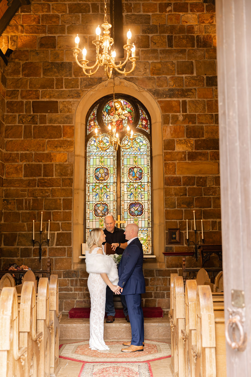 A couple getting married in a church, standing before a stained glass window. They're holding hands, with a chandelier overhead.