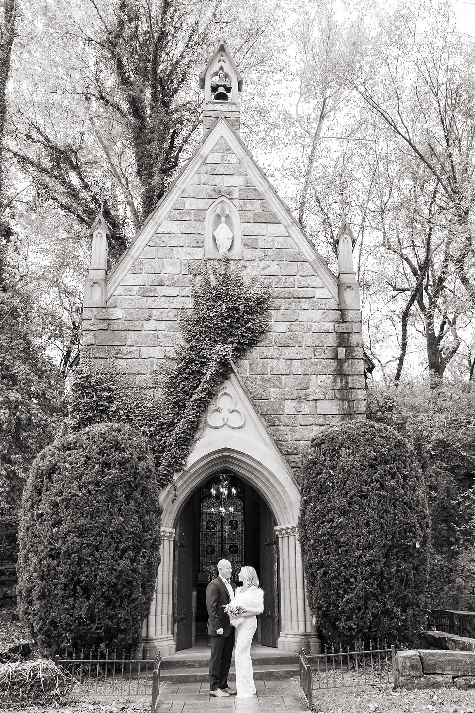 Couple stands smiling in front of an ivy-covered stone chapel. The setting is serene with tall trees surrounding the structure. Black and white.