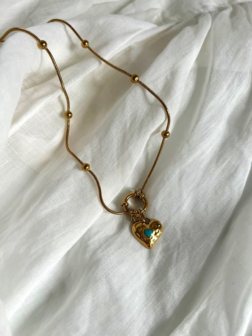 Goldene Halskette mit Herzanhänger und türkisfarbenem Stein auf weißem Stoff.