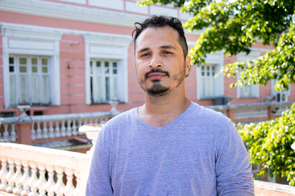 Usando uma blusa lilás de manga comprida, Pedro Martins está de pé e sério na foto. Ele é um homem pardo e possui olhos castanhos, cabelos curtos, ondulados e escuros e barba e bigode da mesma cor do cabelo. Atrás dele, há uma árvore do lado direito e um prédio rosa com janelas e mureta de cor branca.