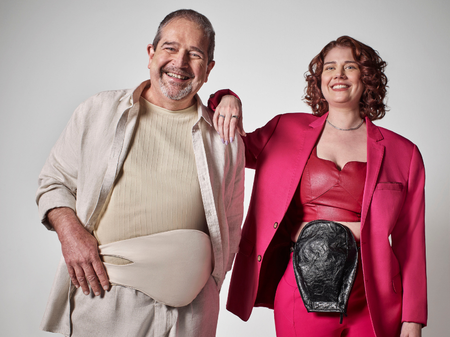 Imagem de um homem e uma mulher sorridentes, em que ele está com a mão direita encostada na alça da bolsa de ostomia bege que usa e ela está com o braço direito apoiado no ombro dele. O modelo masculino usa uma calça e uma blusa bege com uma camisa de manga comprida aberta por cima de mesma cor. Ele é um senhor de meia-idade que possui pele branca, cabelo curto e grisalho e bigode e cavanhaque de mesma cor. Já a modelo feminina usa conjunto de calça e paletó pink com um top de mesma cor, além de uma bolsa de ostomia de cor preta. Ela possui pele branca e cabelos ondulados e ruivos na altura dos ombros. O fundo da imagem é de cor neutra.