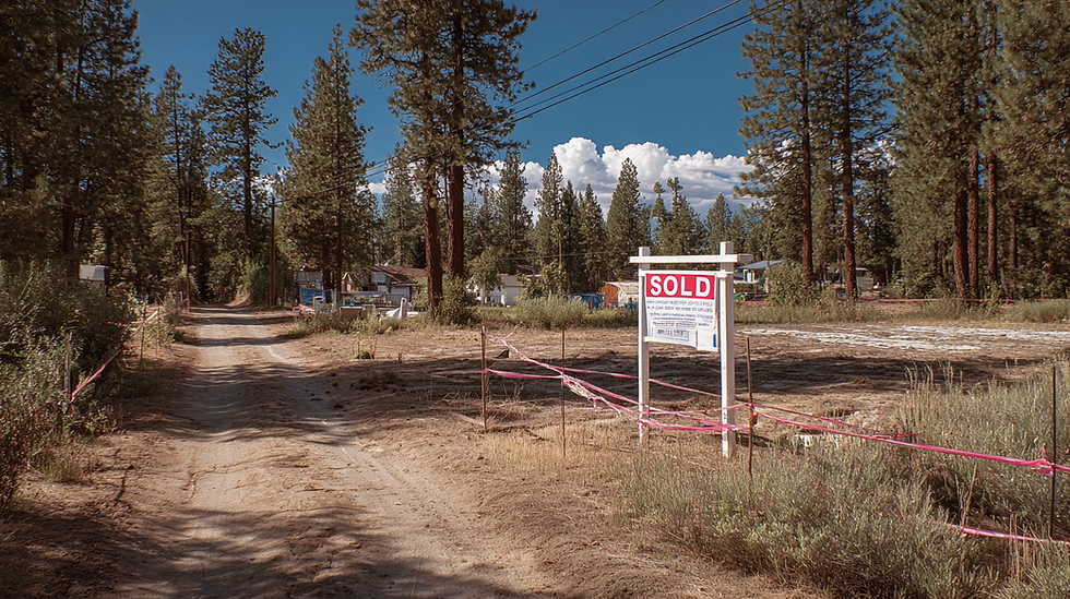 a vacant lot in a pine forest on a dirt road with power lines along it, the corners of the lot are marked with stakes and ribbons, there is a large for sale sign on the lot facing the road that says "SOLD!" on it