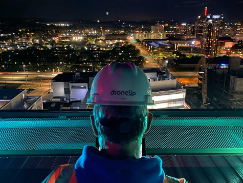 Man in safety helmet standing on the top of a building looking over a colorful city lit up at night.