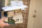 Person holding Amazon packages and envelope by brick wall and door. Boxes show logos and blue Prime tape. Bright daytime setting.