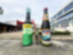 Person joyfully poses between two giant soy sauce bottles outdoors, with a factory and greenery in the background. Bottles display Lee Kum Kee branding.