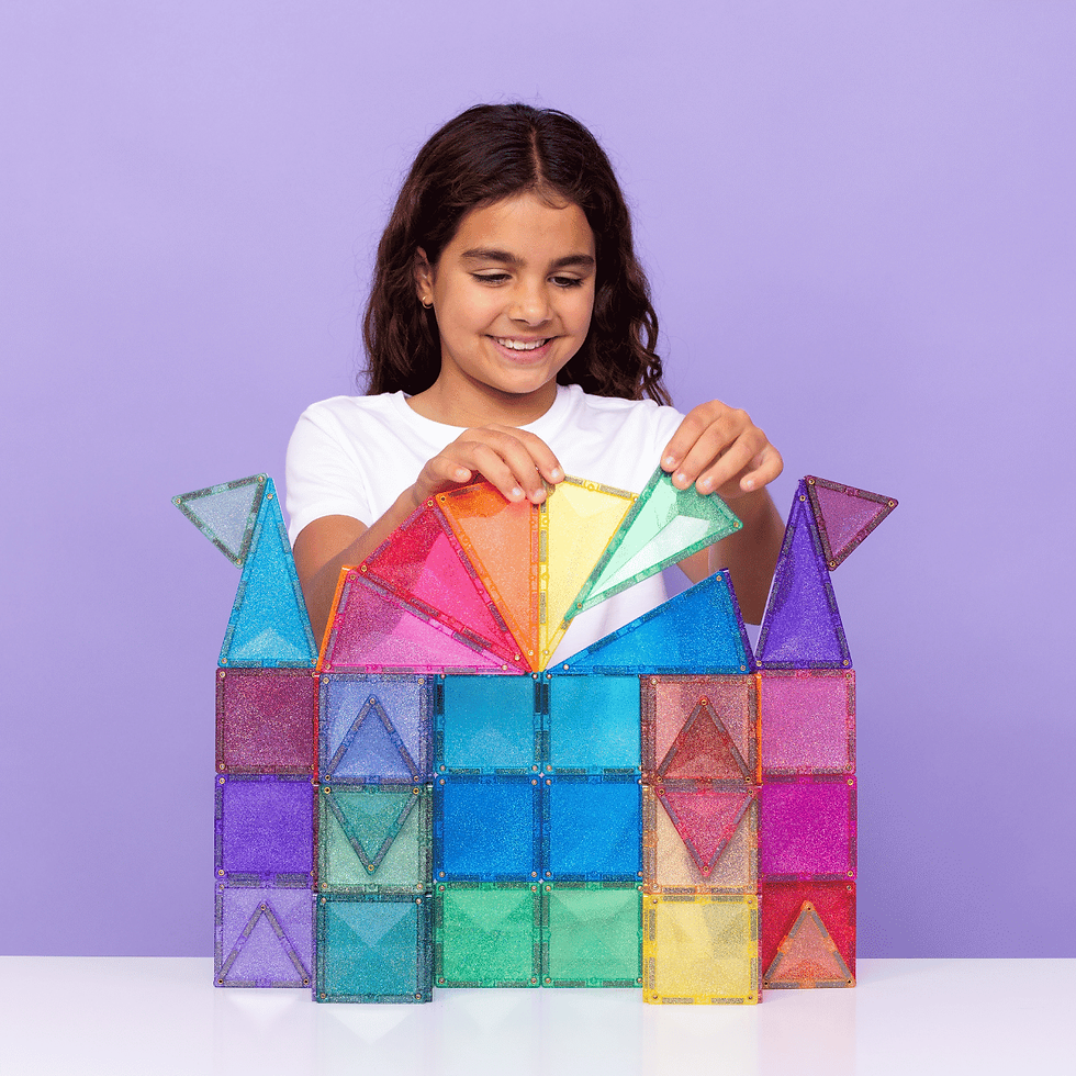 Girl in white shirt smiles while building colorful translucent block structure on table; purple background adds a playful mood.