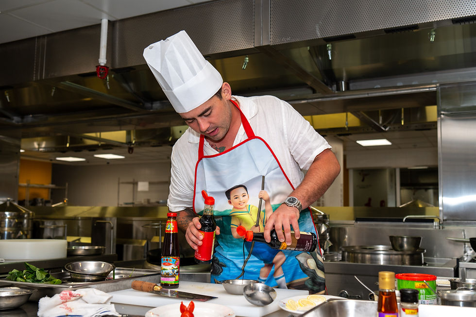 Chef in white hat and colorful apron pours sauce in a commercial kitchen. Stainless steel background, vibrant bottle labels visible.
