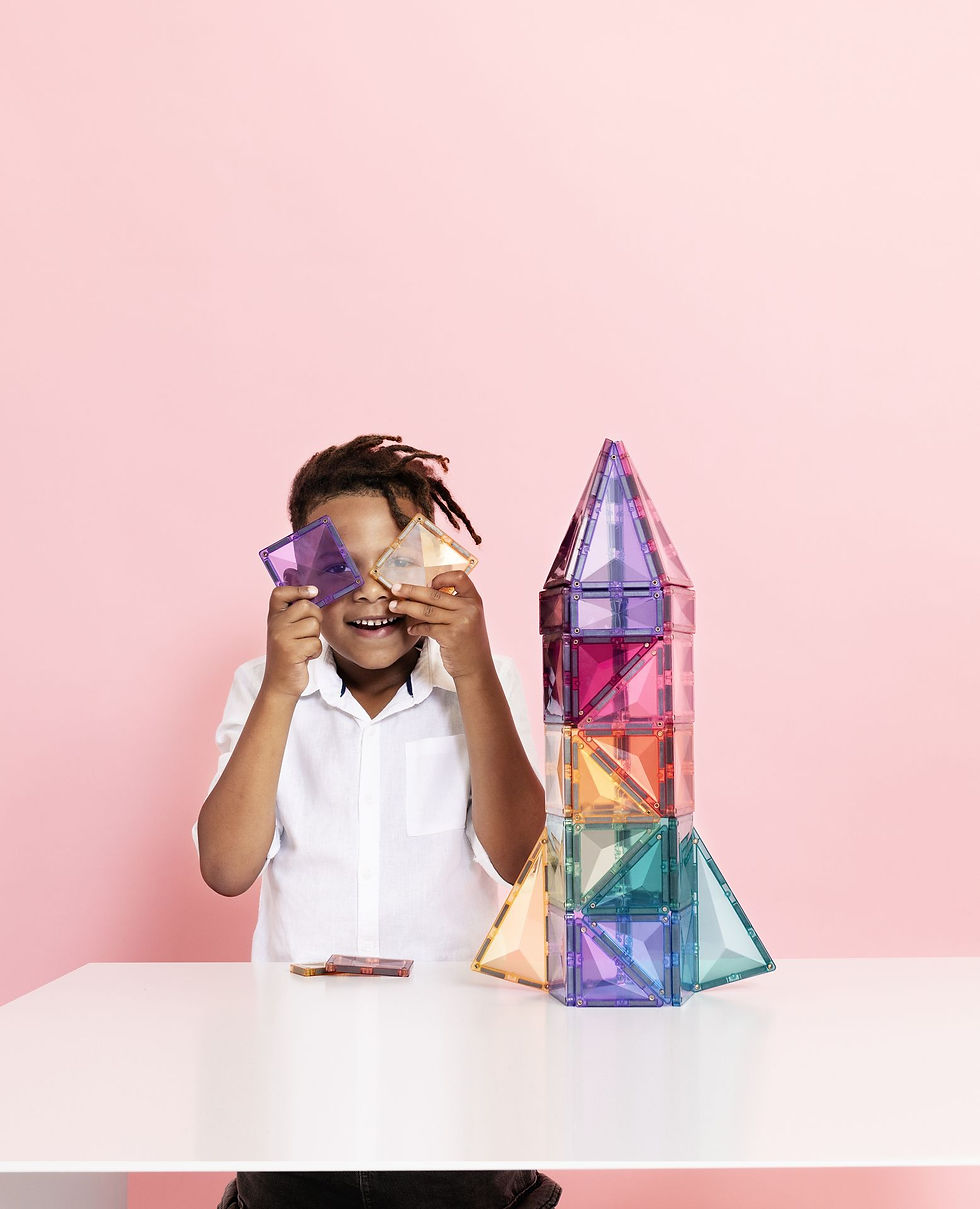 Child in a white shirt holds colorful magnetic tiles to their eyes, smiling, beside a rocket-shaped tile structure on a table. Pink background.