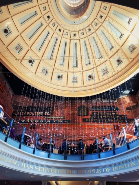 Dome ceiling to Quincy Market building with balcony showing tables and chairs and people seated. Red brick wall with old fashioned decor.