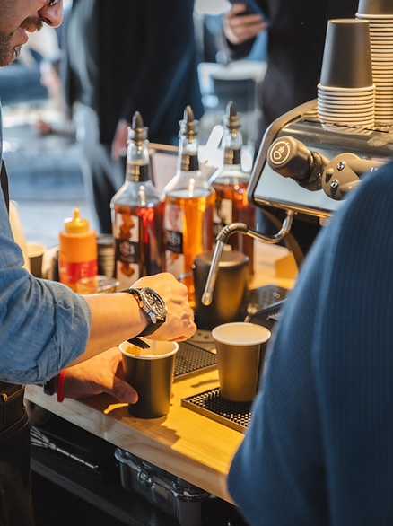Barista pouring coffee, using espresso machine, with syrup bottles in background.