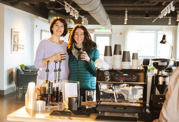 Two women smiling with coffee beside an espresso machine.