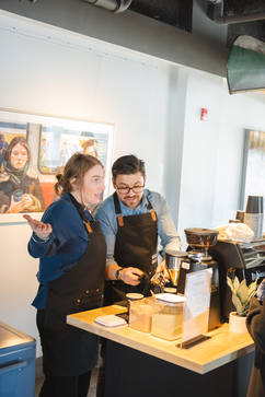 Two baristas working together behind a coffee counter with an espresso machine.