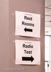 Signs on a column with "Rest Rooms" and an arrow pointing to the left while a sign "Radio Test" with an arrow pointing to the right.