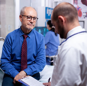 An older man sits on a consultation bed and listens to the doctor explain his test results