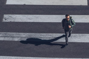 Man in green jacket and backpack takes a short walking break across a zebra crossing. Bright sunlight casts a long shadow on the asphalt.