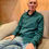 Thumbnail: men sitting indoors against colourful wall wearing relaxed looking turquoise traditional mens shirt in nettle fabric.