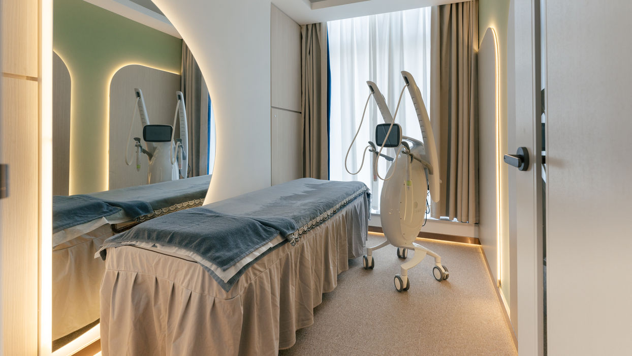 Modern beauty treatment room with a padded bed and contouring machine near floor-to-ceiling curtains.