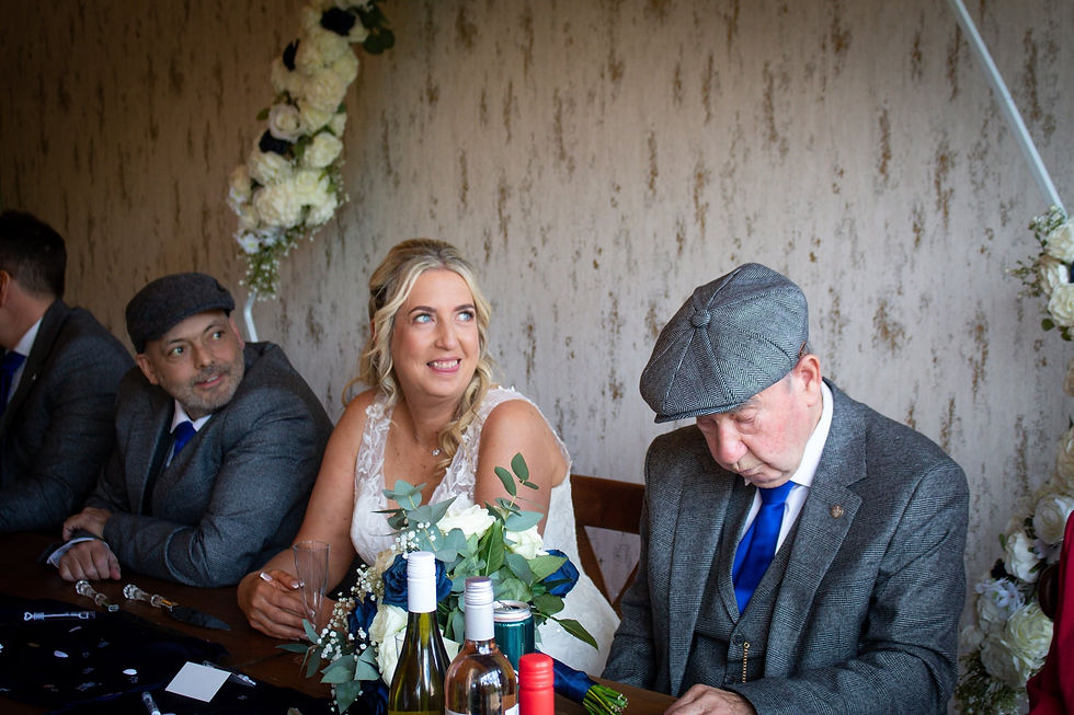 A woman in a white dress smiles beside two men in gray suits and caps. Wine bottles are on the table. Floral arch in the background.