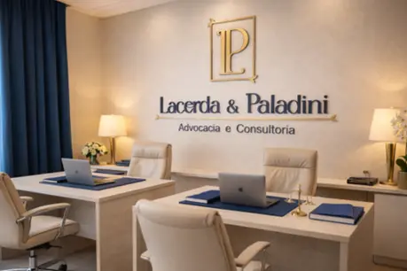 Projeção do ambiente principal de atendimento do escritório, composto por duas bancadas de trabalho em madeira clara dispostas lado a lado com cadeiras executivas bege. Ao fundo, a parede de tom neutro traz a aplicação física do logotipo em dourado e azul-marinho, sob iluminação aconchegante. Cortinas azul-marinho, notebooks, organizadores de mesa e arranjos florais completam o cenário elegante e profissional.