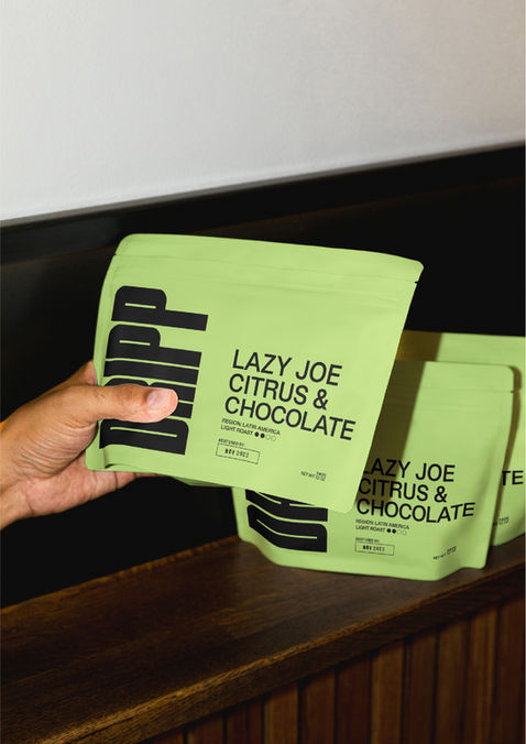 A hand holding a green DRIPP coffee bag. The coffee is called Lazy Joe. There are two other bags in the background on a dark wood table top.