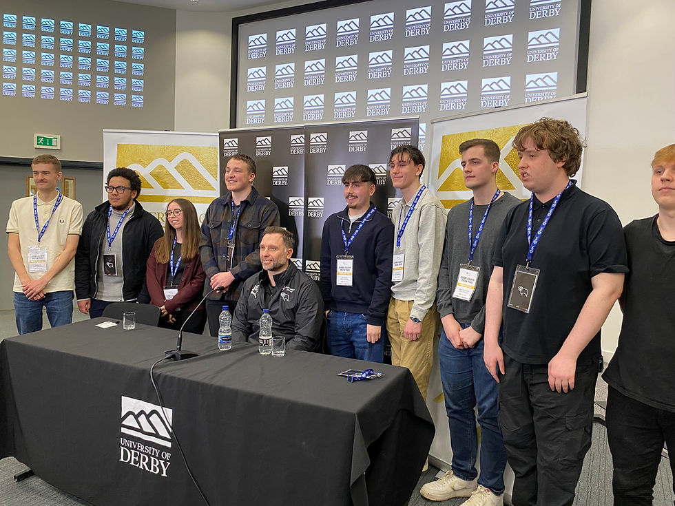 A group of students stand smiling beside a seated John Eustace at a University of Derby press table. Background features a branded screen display.