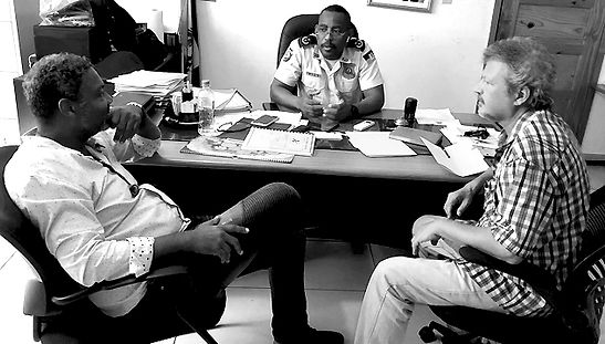 Photo noir&blanc présentant trois hommes dont un policier, il s'agit du Commissaire Jean Gospel. Cette réunion a eu lieu à Petionville Haïti dans le cadre du projet soutenu par l'ONG Periferia Community intitulé "Le Sport au Service de la Paix et du Développement."