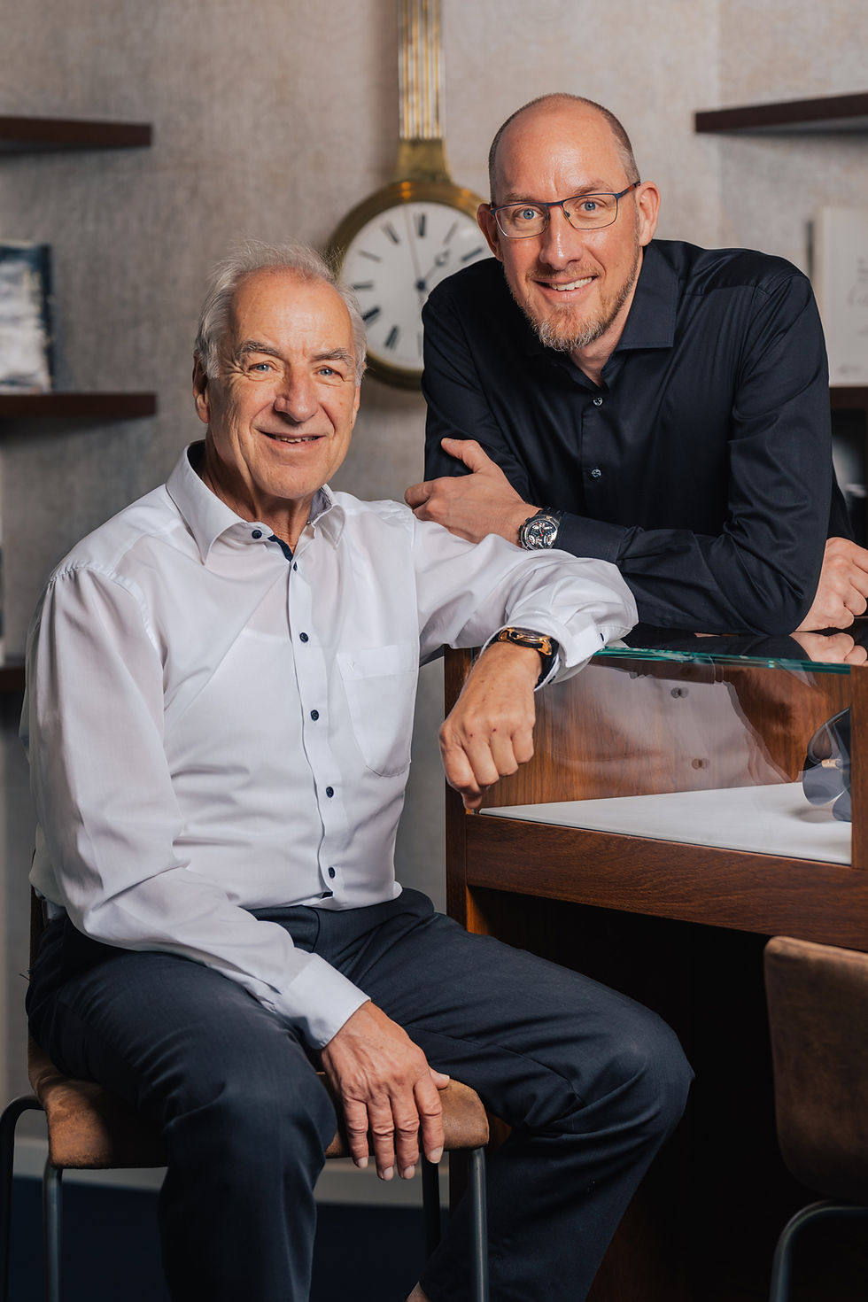 Inhaber und Senior eines Schweizer Uhrenfachgeschäfts an der Verkaufstheke mit antiker Pendeluhr im Hintergrund